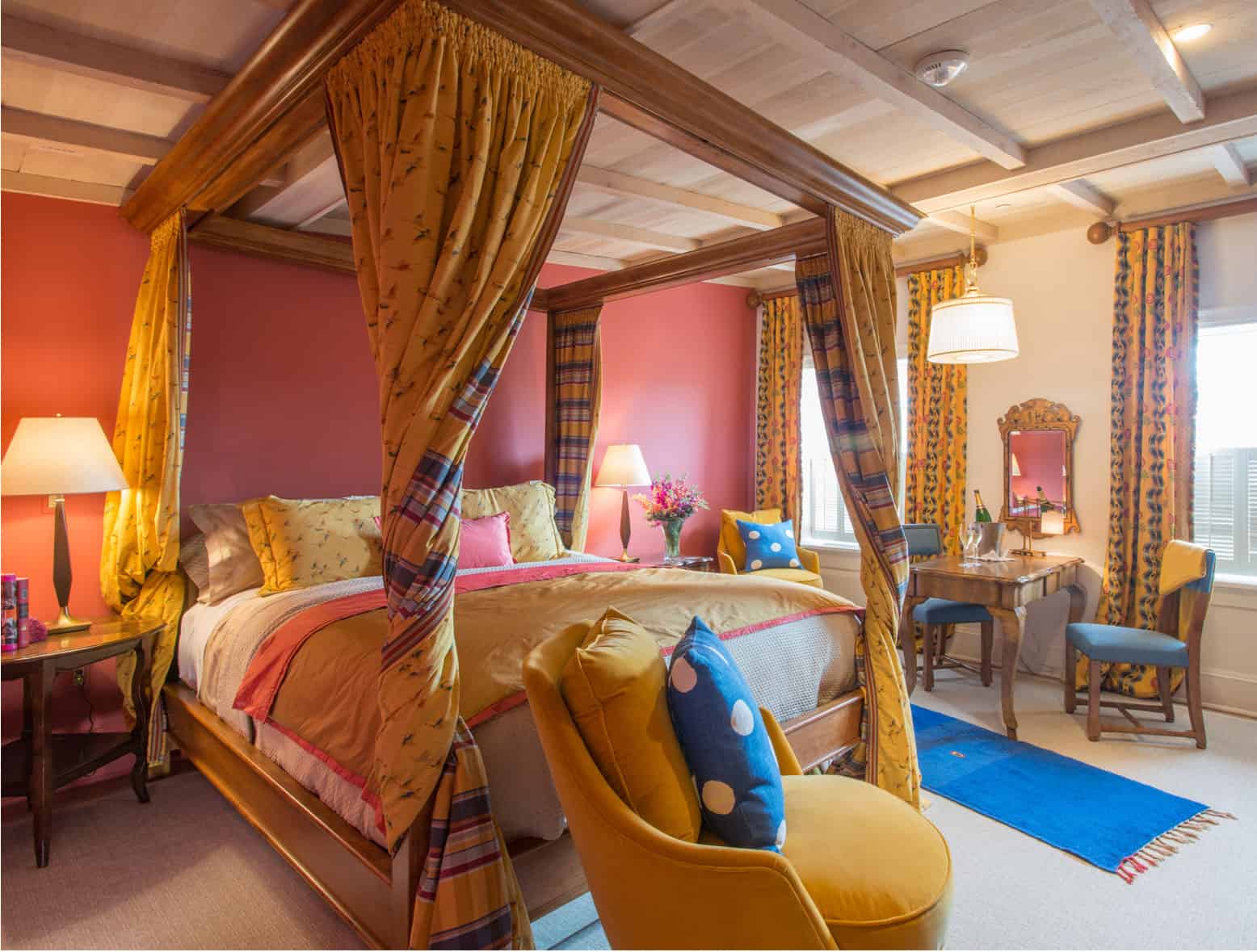 Front perspective view of the four poster bed inside the Ivy Hotel Baltimore. The bedroom goes for a storybook theme, with golds and a pattern for the canopy drapes. A contrasting blue pillow rests on the chair in front