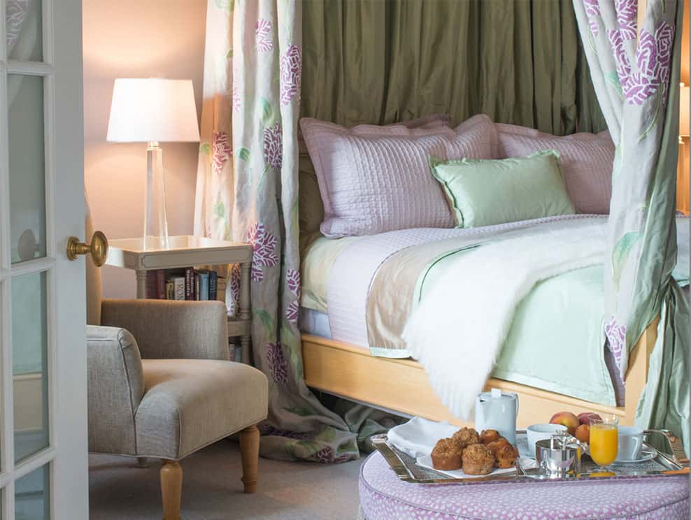 Perspective shot from the left side of the king sized bed. This four poster bed is dressed in pink and light green pastels, with matching drapes that can be drawn for extra privacy. In front, a serving tray of breakfast is set upon the Ottoman.