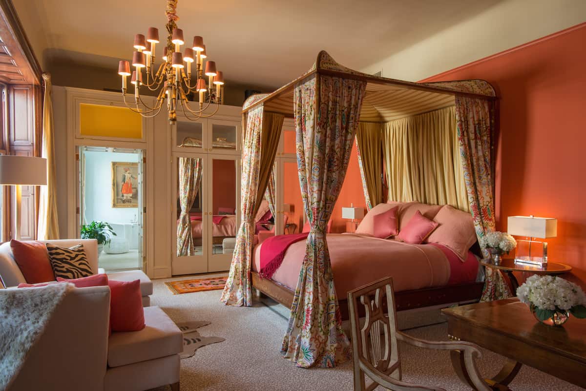 Bedroom of Suite Seven in the Ivy Hotel Baltimore. The four poster king sized bed is placed between two nightshades. This part of the mansion gives a well lit window view, with panel mirrors on the door just beyond.