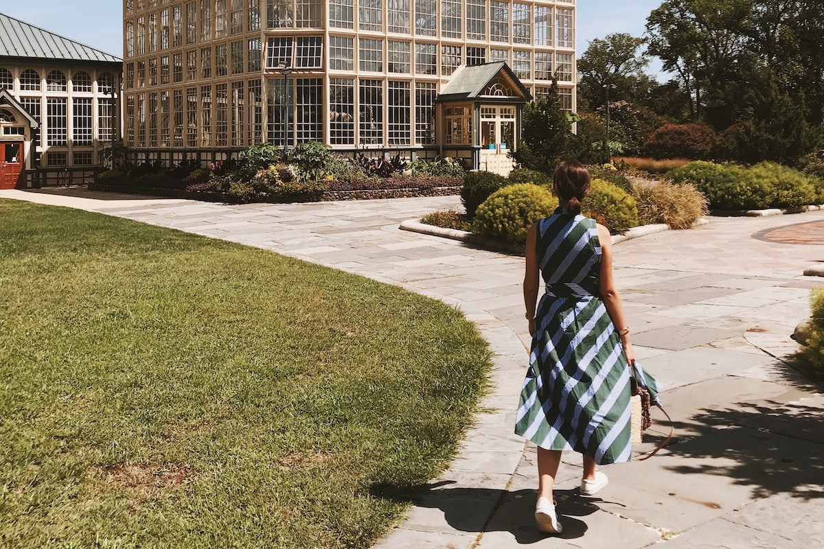 Woman in blue and green striped dress walking outside on a stone path toward a conservatory.