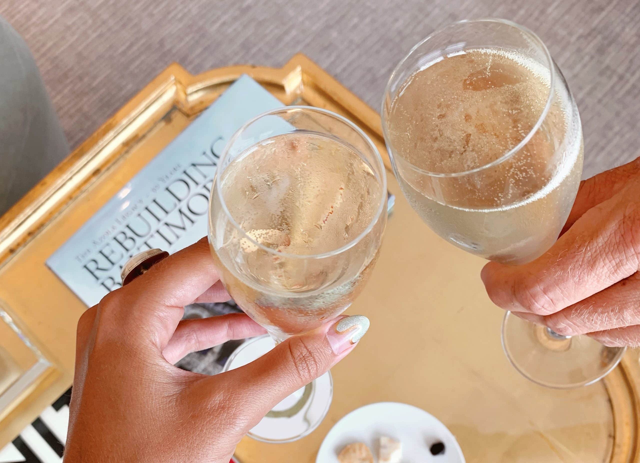 two hands doing a toast with glasses of champagne over a coffee table with treats on it at The Ivy Hotel.